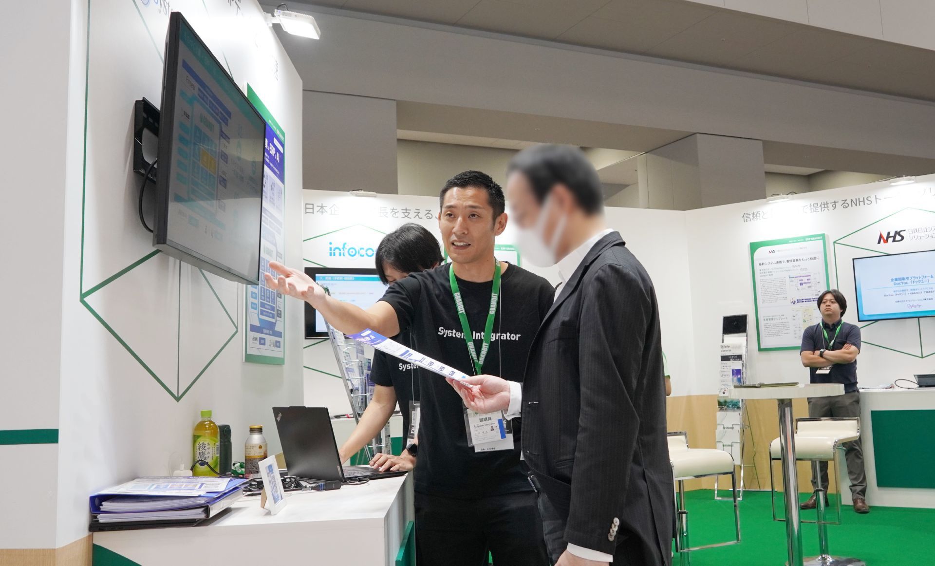 展示会の株式会社システムインテグレータのブースで、来場者に製品の説明をするスタッフの様子